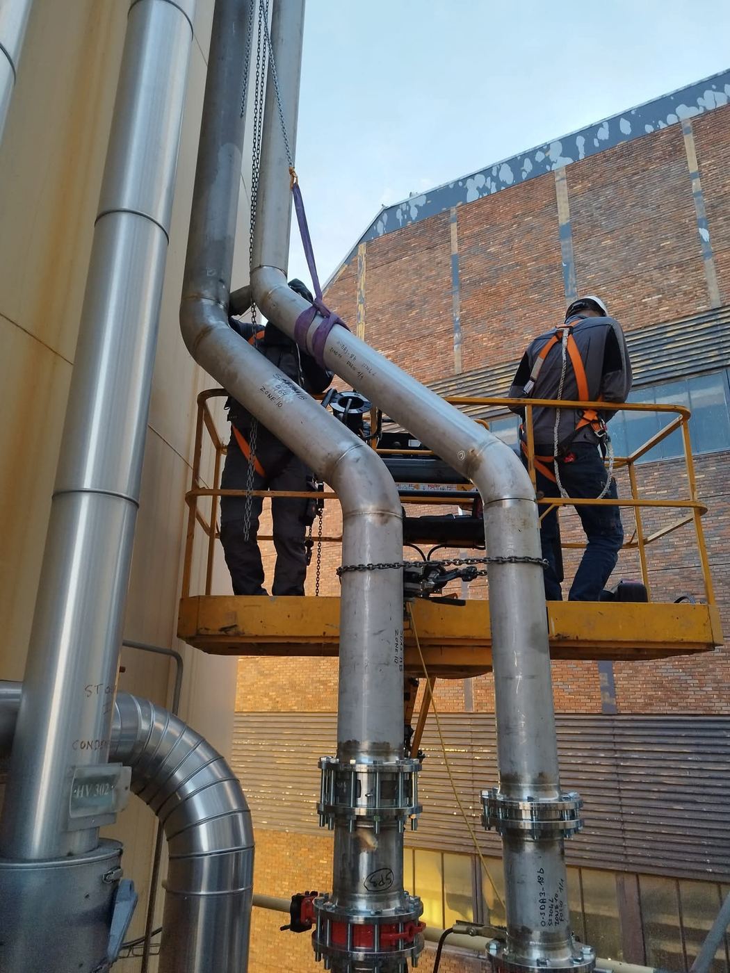 Deux compagnons en nacelle raccordent des tuyauteries inox le long d'un réservoir